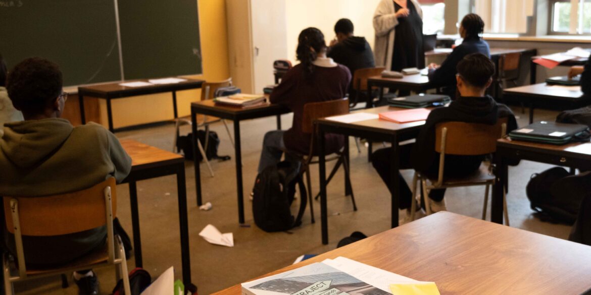 Een klaslokaal, waarbij de juffrouw les geeft aan leerlingen. De focus van de foto ligt op een bureau met schoolboeken