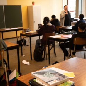 Een klaslokaal, waarbij de juffrouw les geeft aan leerlingen. De focus van de foto ligt op een bureau met schoolboeken