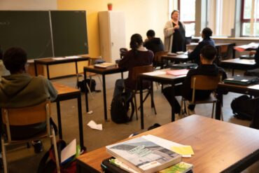 Een klaslokaal, waarbij de juffrouw les geeft aan leerlingen. De focus van de foto ligt op een bureau met schoolboeken