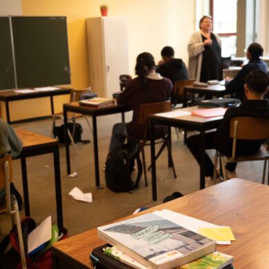 Een klaslokaal, waarbij de juffrouw les geeft aan leerlingen. De focus van de foto ligt op een bureau met schoolboeken