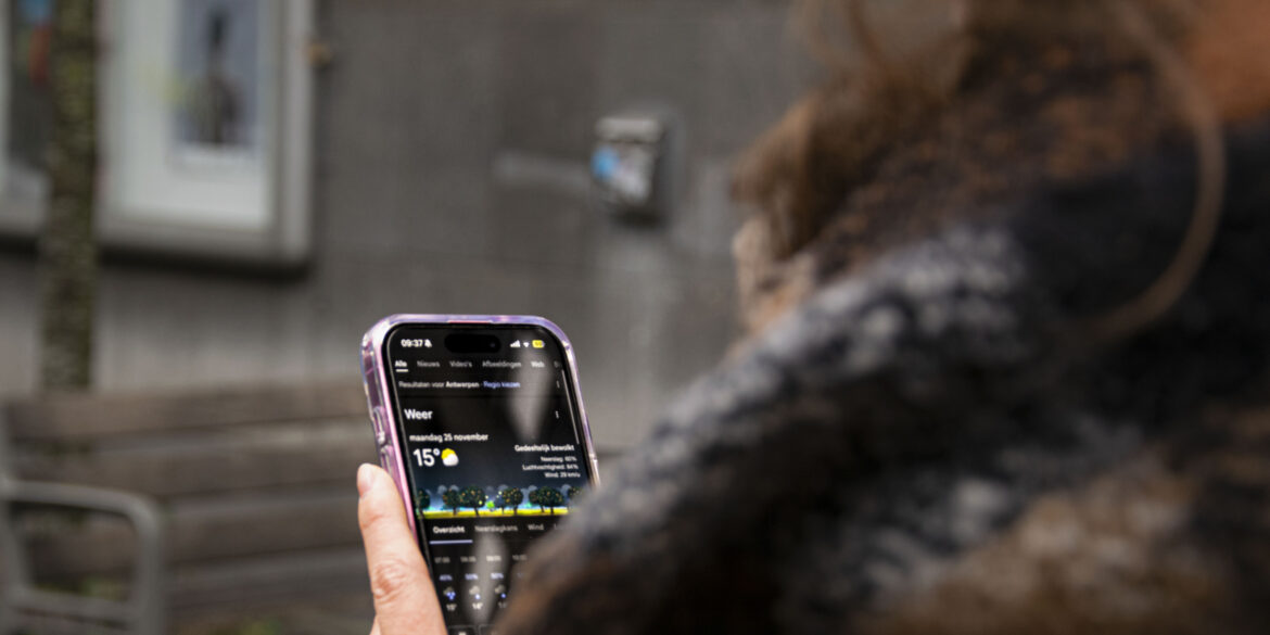 Vrouw met sjaal aan die op haar gsm naar het weerbericht van dit weekend kijkt. Het zegt 15 graden.