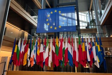 Display of European Union member state flags in front of a large EU flag inside a modern, glass-and-steel architectural building with natural light streaming in.